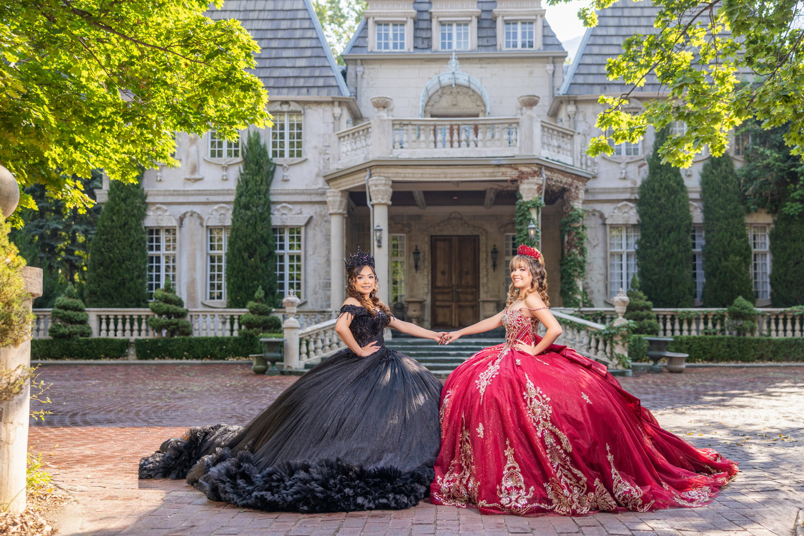 Dos quinceañeras con vestidos negro y rojo tomadas de la mano frente a un edificio de piedra